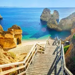 Schody prowadzą na piaszczystą plażę z turkusową wodą, idealne miejsce na portugalskie wakacje. Klify i skały tworzą malowniczy krajobraz.