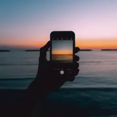 Dłoń trzyma smartfon, na którym widać piękny wschód i zachód słońca nad morzem.