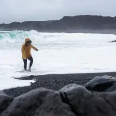 Kobieta w żółtej kurtce brodzi w pianie na czarnej plaży Reynisfjara, podczas gdy potężne fale rozbijają się o brzeg.