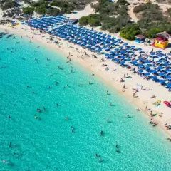 Słoneczny dzień na plaży Makronissos. Turkusowa woda, biały piasek i mnóstwo niebieskich parasoli. Ludzie kąpią się i opalają.