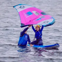 Dziecko z instruktorem na desce, trzymają różową żagiel do wingfoil. Radość na twarzy dziecka.
