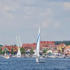 Żaglówki pływają po błękitnej wodzie w malowniczej zatoce, w tle widać urokliwe miasteczko z czerwonymi dachami i wieżą kościelną.