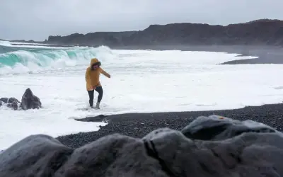 Kobieta w żółtej kurtce brodzi w pianie na czarnej plaży Reynisfjara, podczas gdy potężne fale rozbijają się o brzeg.