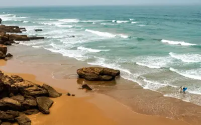 Maroko morze. Dwie osoby z deskami surfingowymi wchodzą do wody na piaszczystej plaży, fale rozbijają się o skaliste wybrzeże.