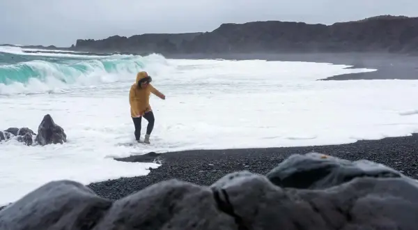 Kobieta w żółtej kurtce brodzi w pianie na czarnej plaży Reynisfjara, podczas gdy potężne fale rozbijają się o brzeg.
