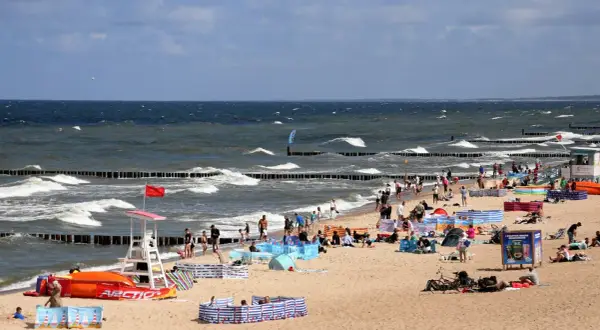 Plaża nad Bałtykiem pełna ludzi, fal i parawanów. Widać ratownika, sprzęt wodny i tablicę informacyjną. Ceny nad morzem pewnie wysokie, ale widok wart zachodu.