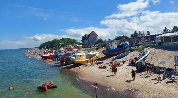 Słoneczny dzień nad polskim morzem. Kolorowe łodzie na plaży, tłumy ludzi, a na wodzie skuter wodny.