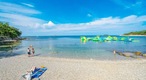 Słoneczna plaża z kamykami, ludzie kąpią się w krystalicznie czystej wodzie, a w oddali wodny plac zabaw. Idealne miejsce na wakacje w camping Bijela Uvala.