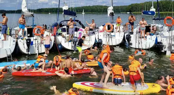 Ludzie w kamizelkach ratunkowych i strojach kąpielowych pływają i bawią się na deskach SUP przy żaglówkach. Wspaniały weekend czerwcowy na wodzie.