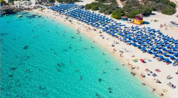 Słoneczny dzień na plaży Makronissos. Turkusowa woda, biały piasek i mnóstwo niebieskich parasoli. Ludzie kąpią się i opalają.
