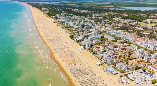 Słoneczny dzień nad morzem. Tłumy plażowiczów na szerokiej, piaszczystej plaży, a za nią miasteczko z hotelami i zielenią.