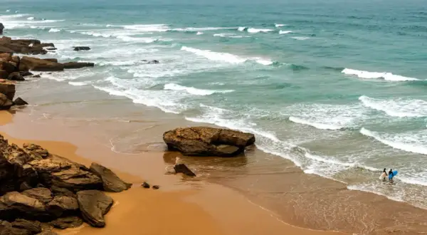Maroko morze. Dwie osoby z deskami surfingowymi wchodzą do wody na piaszczystej plaży, fale rozbijają się o skaliste wybrzeże.