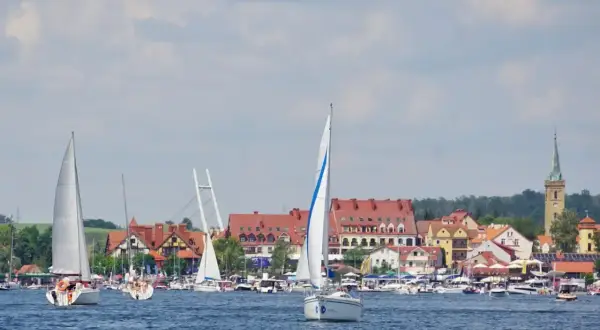 Żaglówki pływają po błękitnej wodzie w malowniczej zatoce, w tle widać urokliwe miasteczko z czerwonymi dachami i wieżą kościelną.