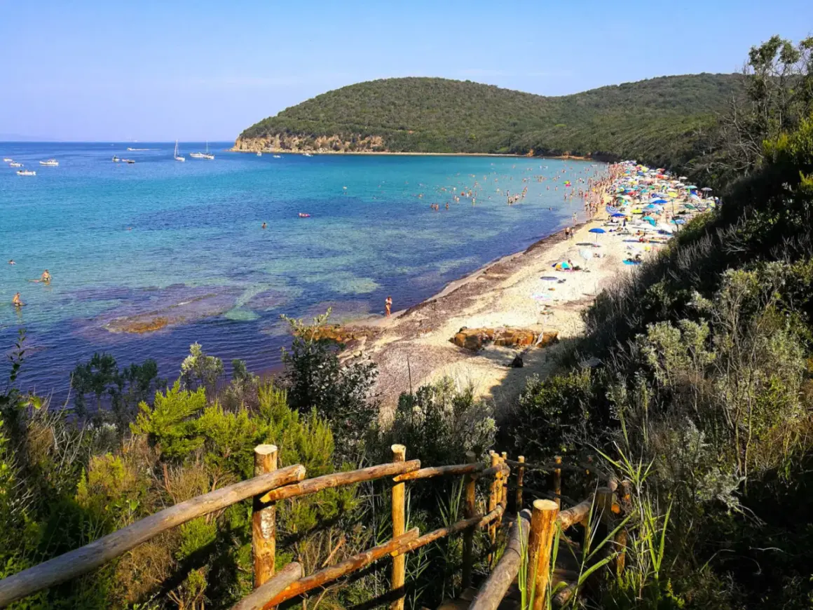 Słoneczne livorno plaże pełne ludzi, z turkusową wodą i zielonymi wzgórzami w tle. Drewniane schody prowadzą na piaszczysty brzeg.