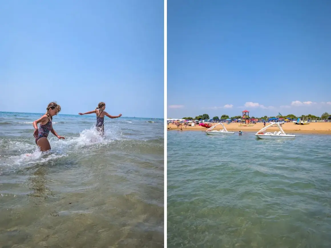 Dwie dziewczynki pluskają się w wodzie na plaży Bibione. W tle widać łódki i parasole.