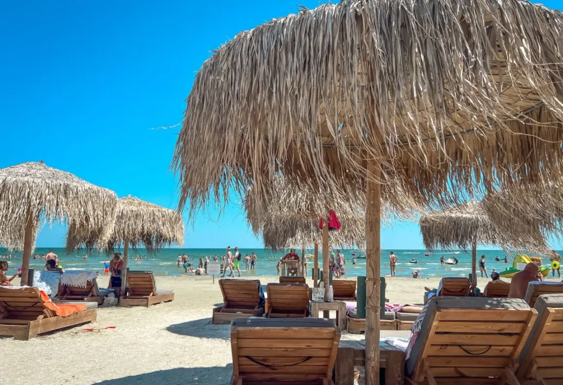 Słoneczne wakacje w Rumunii nad Morzem Czarnym. Leżaki i parasole z trawy na piaszczystej plaży, w tle kąpielowicze.