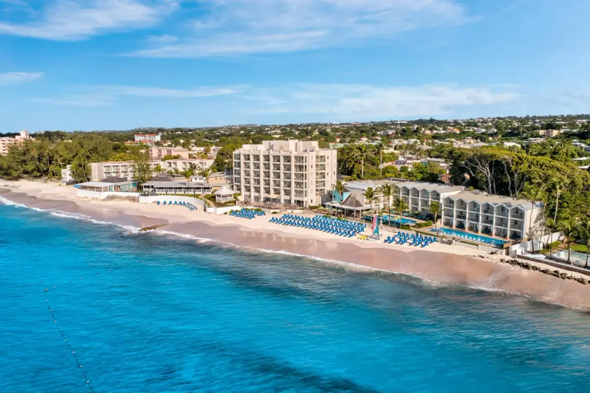 Idealne Barbados wakacje: hotel przy plaży z basenami i leżakami, turkusowe morze i zielone wzgórza w tle.