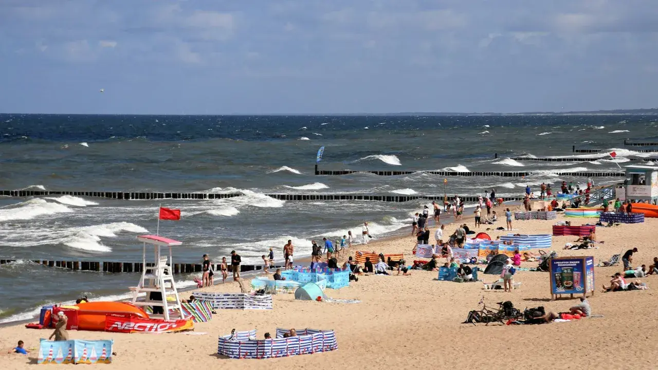Plaża nad Bałtykiem pełna ludzi, fal i parawanów. Widać ratownika, sprzęt wodny i tablicę informacyjną. Ceny nad morzem pewnie wysokie, ale widok wart zachodu.