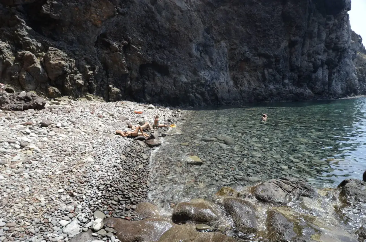 Kamieniste livorno plaże, gdzie słońce ogrzewa kamienie, a woda kusi do kąpieli. Kilka osób odpoczywa na brzegu, jedna pływa w krystalicznie czystej wodzie.