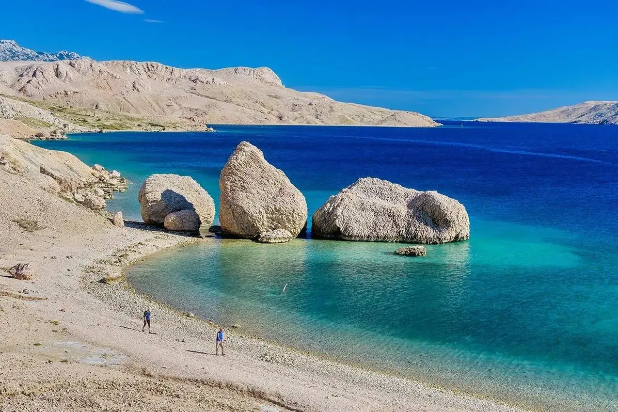 Kamienista plaża na wyspie Pag z turkusową wodą i skalistymi górami w tle. Dwie osoby spacerują brzegiem.